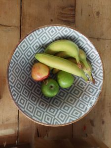 A geometric-patterned bowl sits on a wooden table, holding several pieces of fruit including three bananas, a red-and-yellow apple, a green apple, and a green citrus fruit.