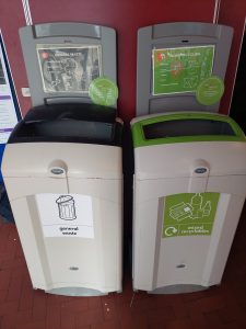 wo indoor waste bins placed side by side against a red wall: one labeled “general waste” with a black lid, and the other labeled “mixed recycling” with a green lid. Each bin has a sign above it showing examples of what can be disposed inside, along with a circular notice advising not to throw liquids in the bins.