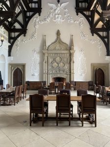 A hall with ornate plasterwork on the walls and a beamed ceiling. There are several wooden tables and chairs set out for dining. 