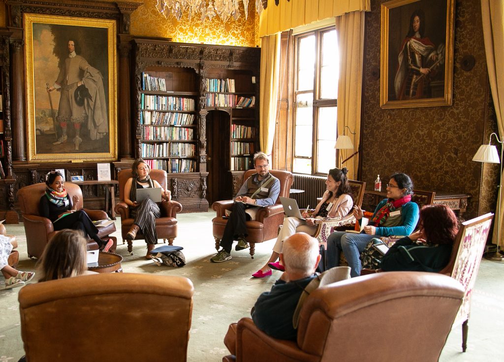 A group of people sitting in armchairs in a circle. There are bookshelves and two grand paintings behind them. 