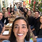 Large group seated at long outdoor table, sharing pizza and drinks at a sunny café with plants and busy background.