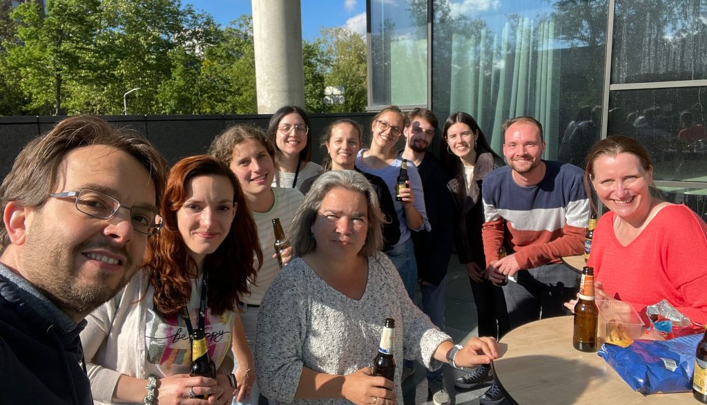 A lab group posing for a selfie with drinks. They are sitting outside a glass building around some tables.