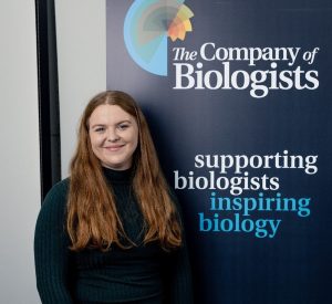 Erin Brown posing for a photograph in front The Company of Biologists and the slogan "supporting biologists inspiring biology". 
