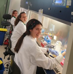 Three scientists wearing lab coats and sitting at fume hoods, where they are pipetting cells. 