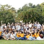 A large, diverse group of young people posing together outdoors on a grassy lawn, smiling, cheering, and raising their arms, with trees and greenery in the background