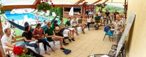 Adam Kampff stands in front of a group of seated students, using a whiteboard to deliver a lecture to them. A swimming pool is visible in the background behind the students. 