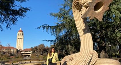 A person standing in front of a worm-shaped sculpture. A university campus building can be seen in the background