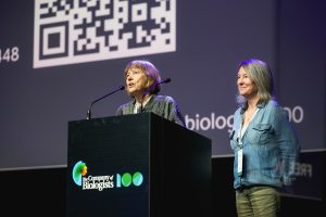 Two women stand on a stage behind a podium labeled The Company of Biologists. One woman speaks into a microphone, while the other stands beside her, both facing the audience. A large screen with a QR code is visible behind them.
