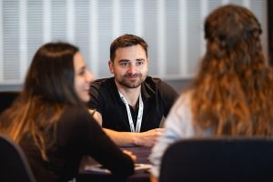 A man wearing a lanyard sits at a table, smiling and engaging in conversation with two women, both with long hair, in a bright indoor setting.