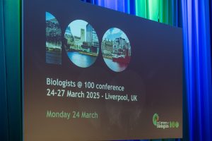 A large screen displays information for the Biologists at 100 conference, taking place 24-27 March 2025 in Liverpool, UK. The background shows city images and green, blue, and black draped curtains.