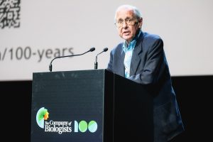 A man in a suit speaks at a black podium with The Company of Biologists 100 logo, against a backdrop with partial text and a QR code. Two microphones are attached to the podium.