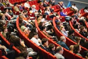 A large audience sits in red theater-style seats, attentively listening to a presentation or lecture. People are of diverse ages and backgrounds, some taking notes, creating a focused and engaged atmosphere.