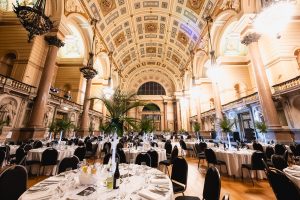 Elegant banquet hall with high, ornate arched ceilings, chandeliers, and large columns. Round tables with white tablecloths, glassware, and centerpieces are arranged for a formal event.